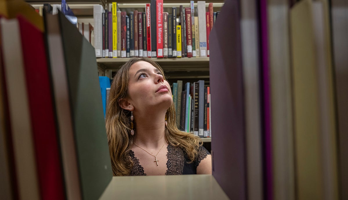 A young woman in a library peers through a shelf of books.