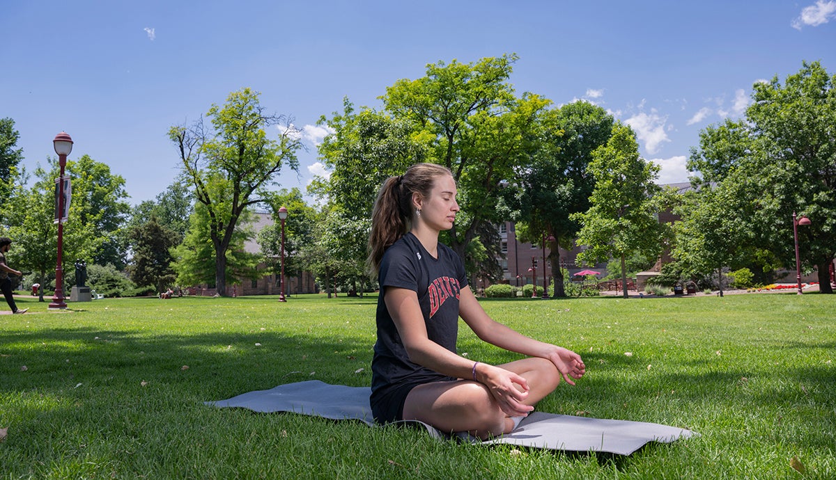 A young woman sits on a yoga mat outdoors with her eyes closed and legs crossed.