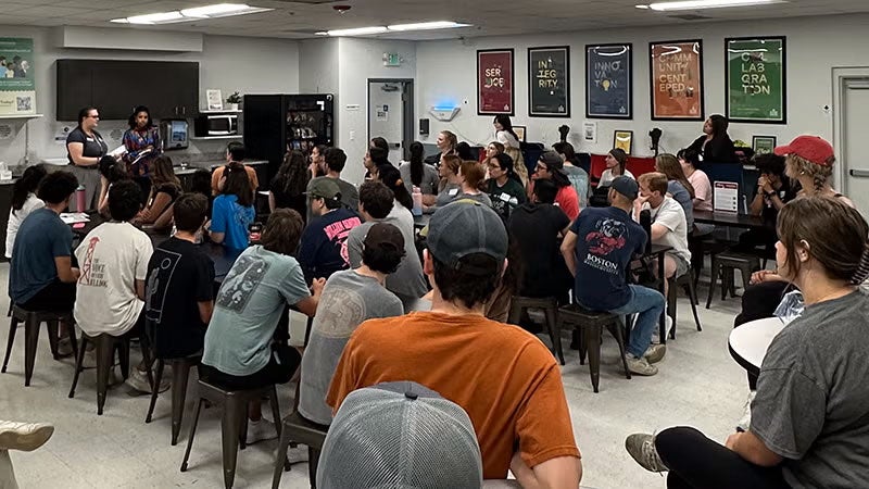 students seated around table in conference room in front of two speakers, standing