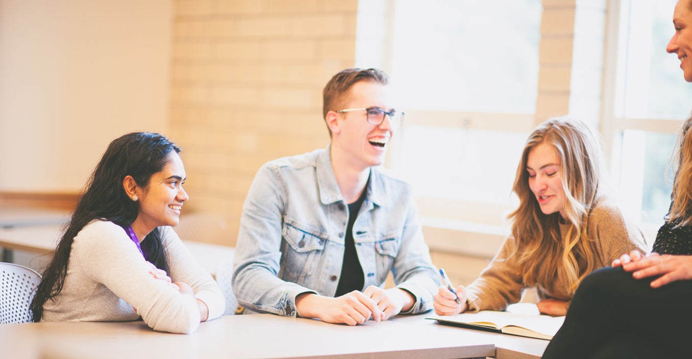 four people smiling at each other. one is writing on her notebook and one is sitting on the desk