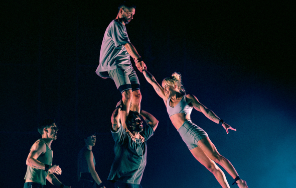 A team of acrobats lifts a woman into the air by one arm.