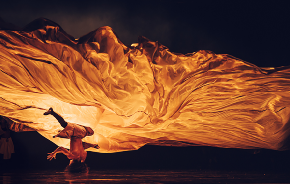 A dancer spins on their head in front of a flowing, golden-lit fabric. 