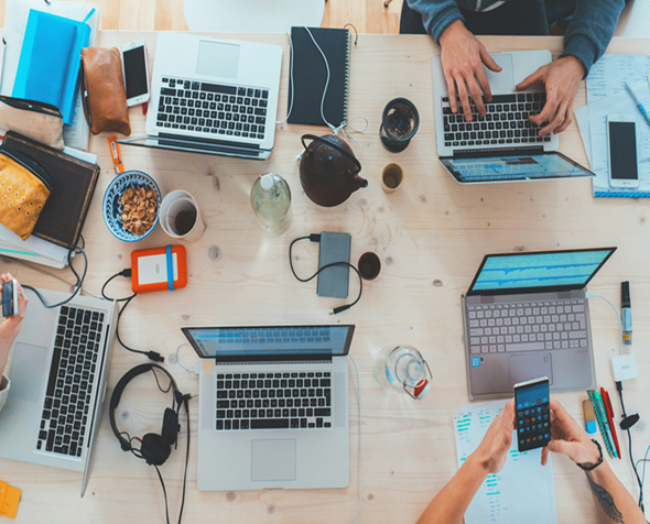Four laptops open on a table with notebooks, phones, hard drives and coffee cups