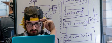 a student staring at his laptop. programming write on the whiteboard behind him
