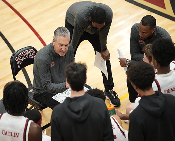 Jeff Wulbrun, seated, clipboard in hand, in huddle with men's basketball players