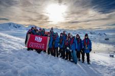 Students huddle for a group photo on the Antarctic Peninsula by Neko Harbor, holding a DU banner.