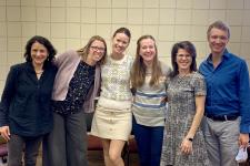 5 people stand with their arms around each others shoulders and smile for their picture to be taken.