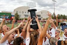 DU women's lacrosse team celebrating around the Big East tournament trophy