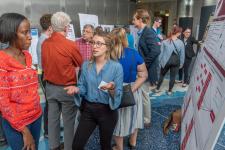 A young woman presents a trifold board research project to several onlookers.