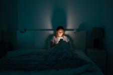 A woman sits alone in a large bed on her phone with the lights off, the light from her phone illuminating her face.