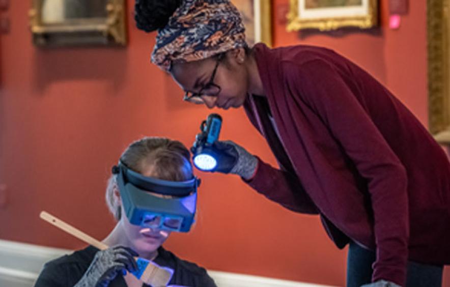 goggle-wearing woman with child holding flashlight as both examine a painting
