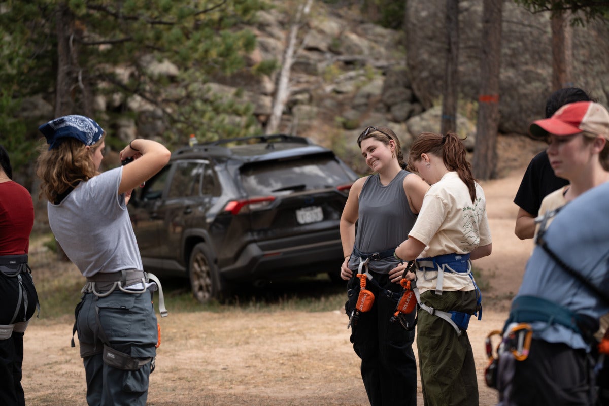 A student in climbing gear takes a picture of her friend, who is also in climbing gear.