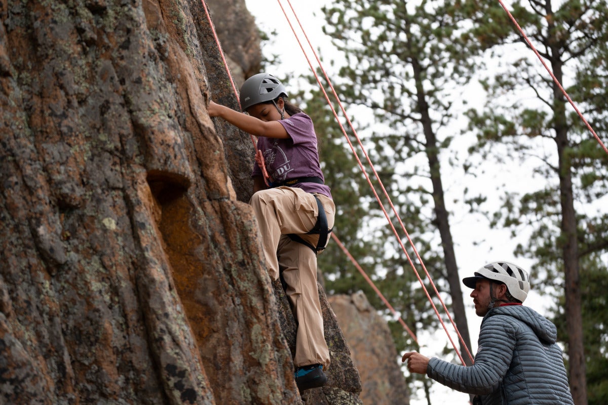 A female student wearing a purple t-shirt, khaki pants, and a helmet scales a cliffside with the help of cables, while her climbing partner observes from below.