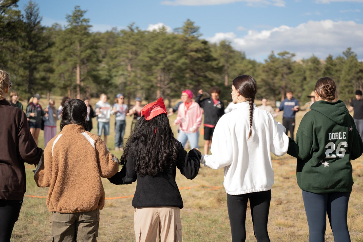 A group of students stands in a circle and holds hands in the field.