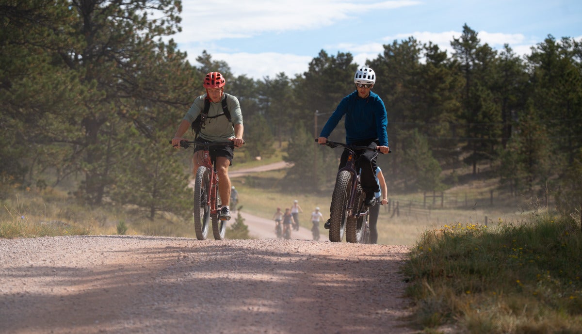 Two male students ride down a dirt road on mountain bikes.