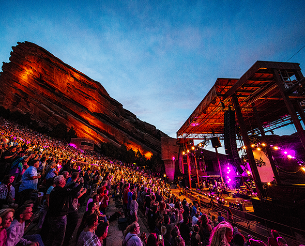 Photo of Red Rocks Amphitheatre at night during a concert with pink lighting on the stage, a large red rock visible in the background and crowded seats in the foreground