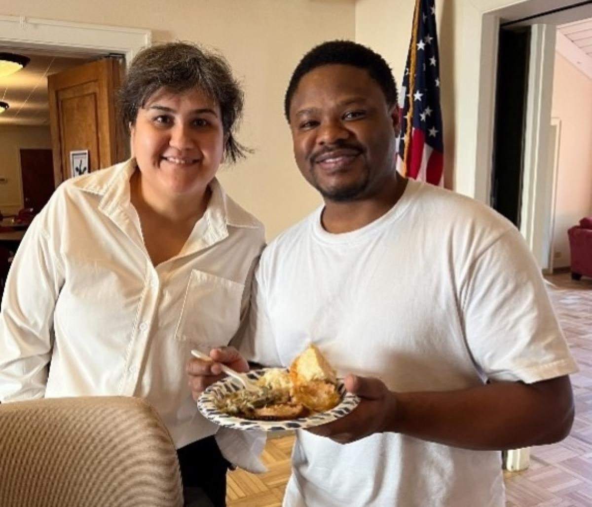 Two people stand together, posing for a photo, with one person holding a plate of food.
