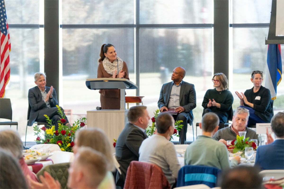 Provost Loboa speaks in front of a group of people, with the Chancellor, Jeff Banks, Abigail Girard and one other woman sit behind her on stage. 