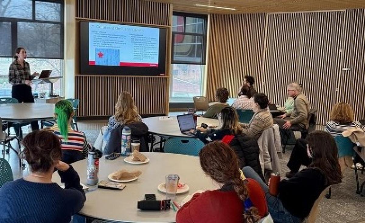 Laura Perille stands behind a podium, speaking to a room full of people while she presents.