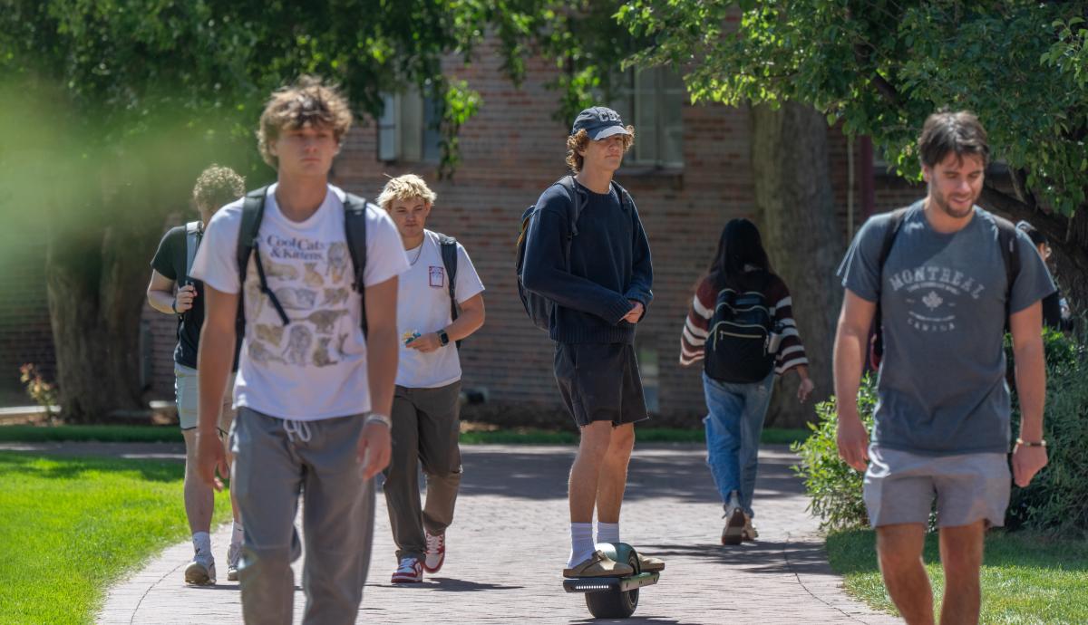 A group of young men walk and skateboard on DU's campus on a nice day.