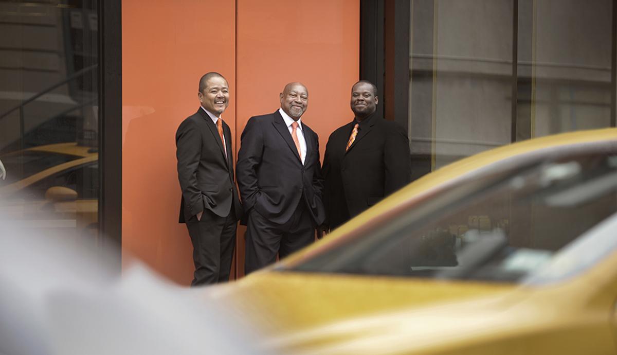 The Kenny Barron Trio stands against a wall with rushing traffic blurred in the foreground.