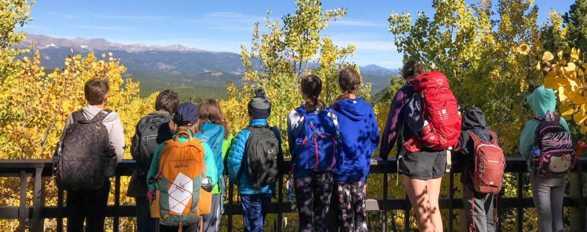 group of children and their supervisor, all wearing backpacks and outdoor gear, viewed from behind as they survey a mountain vista