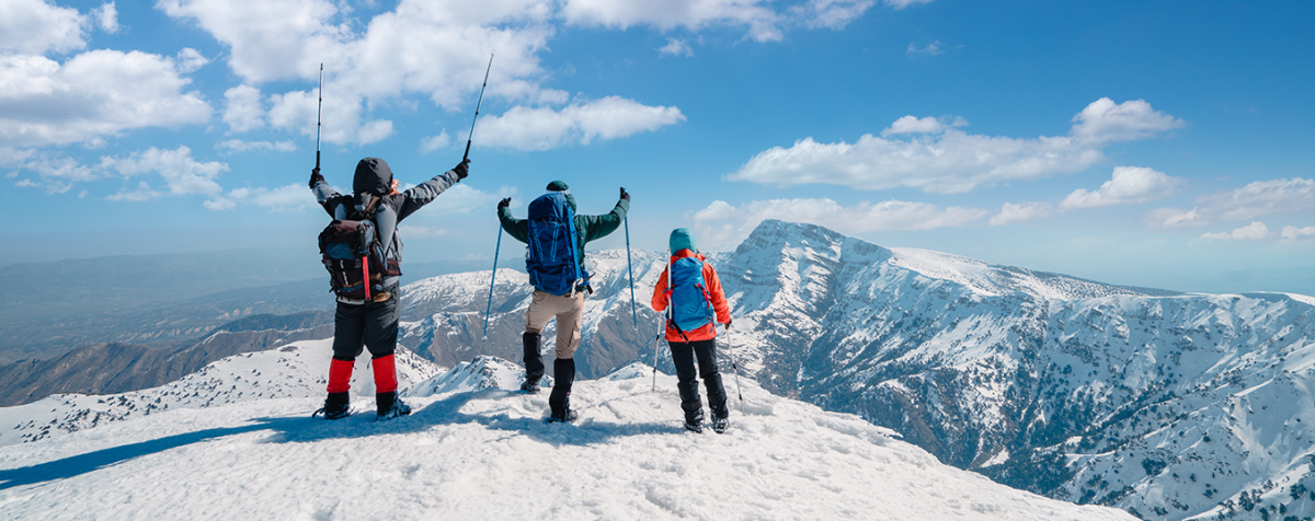 Three skiers on top of a snow covered mountain raise their trekking poles in jubilation.