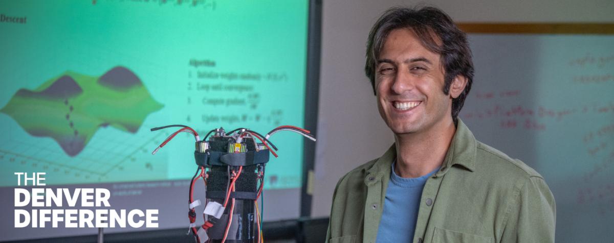 A man smiling with a machine next to him and presentation on the screen on the back