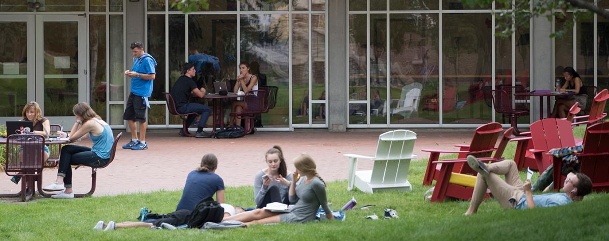 group of students lounging on grass and sitting at tables outside glass-walled building