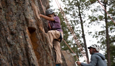 A female student wearing a purple t-shirt, khaki pants, and a helmet scales a cliffside with the help of cables, while her climbing partner observes from below.