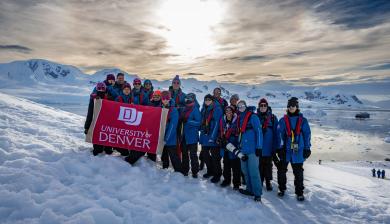 Students huddle for a group photo on the Antarctic Peninsula by Neko Harbor, holding a DU banner.