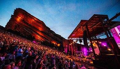 Photo of Red Rocks Amphitheatre at night during a concert with pink lighting on the stage, a large red rock visible in the background and crowded seats in the foreground