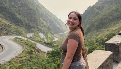 DU student Michela Schenk poses on a bridge overlooking green mountains.