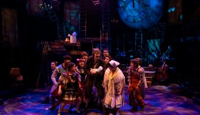 The cast of "Into the Woods" huddles in the center of a colorfully-lit stage. 