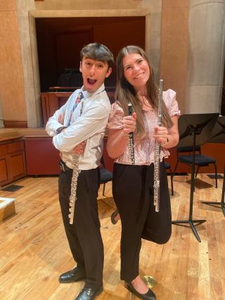 A male and female student in concert attire pose back-to-back, smiling and holding flutes.