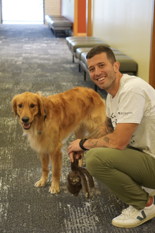 A man in a white t-shirt and green pants crouches down near his golden retriever, holding a dog toy.