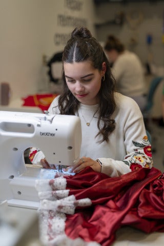 A female student uses a sewing machine to attach white lace to a piece of red fabric.